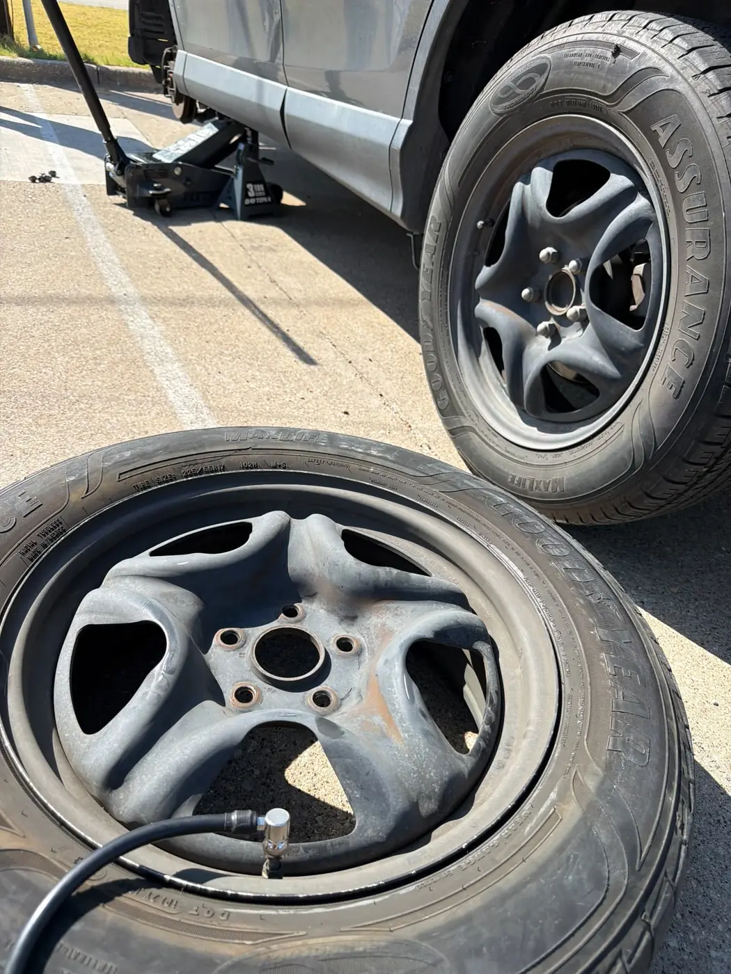 Silver SUV raised on a floor jack and jack stand in a Fort Worth, Texas parking lot with a Goodyear Assurance MaxLife 225/65R17 tire mounted on the alloy and being inflated during an on-site mobile tire replacement.