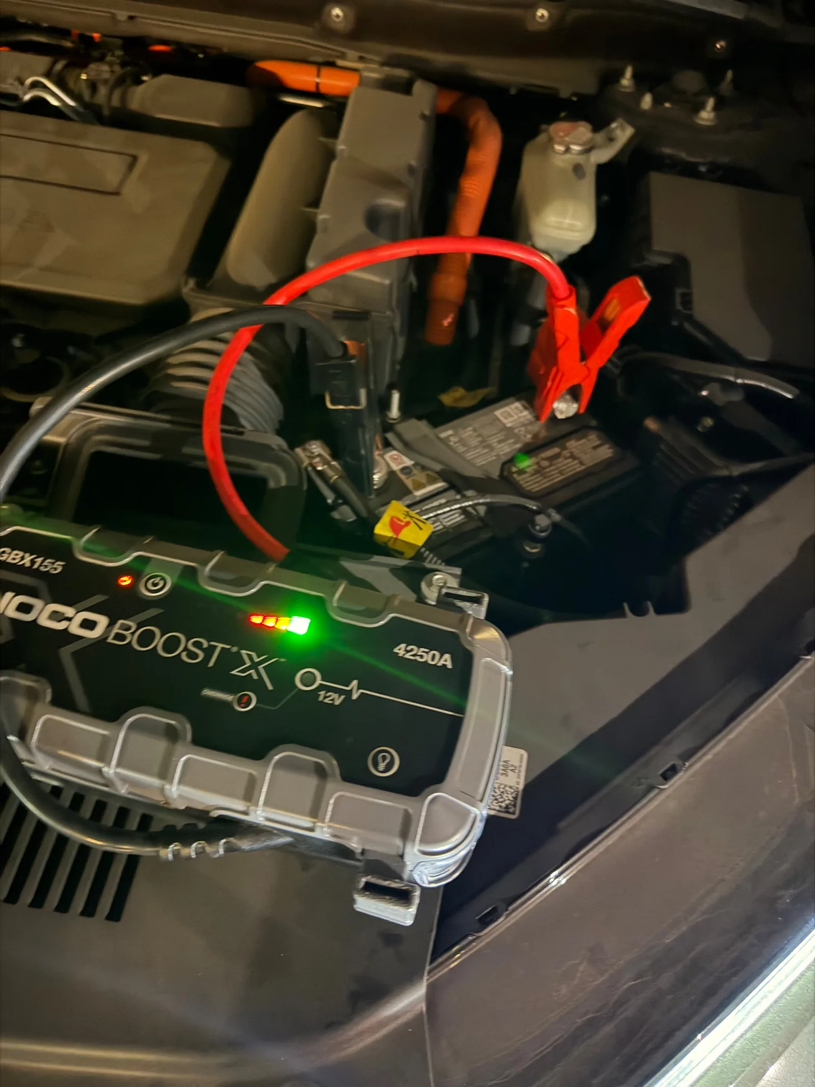 NOCO Boost X GBX155 4250-amp jump pack clamped to the 12V battery of a hybrid vehicle in Southlake, Texas, with the orange high-voltage cabling visible in the engine bay.
