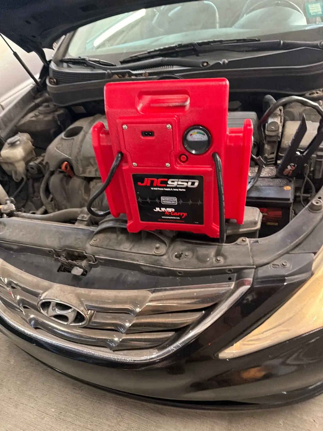 Red JNC950 commercial jump pack resting on the core support of a black Hyundai Sonata with clamps on the battery terminals during a mobile jump start in Dallas-Fort Worth.
