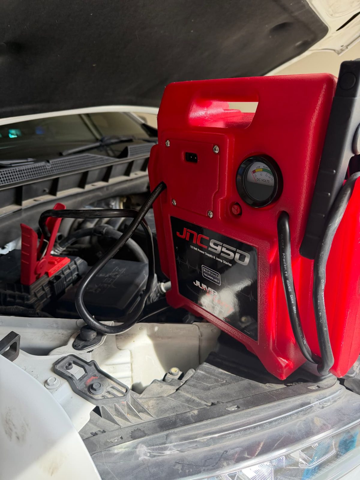 Red JNC950 12-volt jump pack sitting on the radiator support of a white SUV with clamps on the battery terminals during a 24/7 mobile jump start in Dallas-Fort Worth.