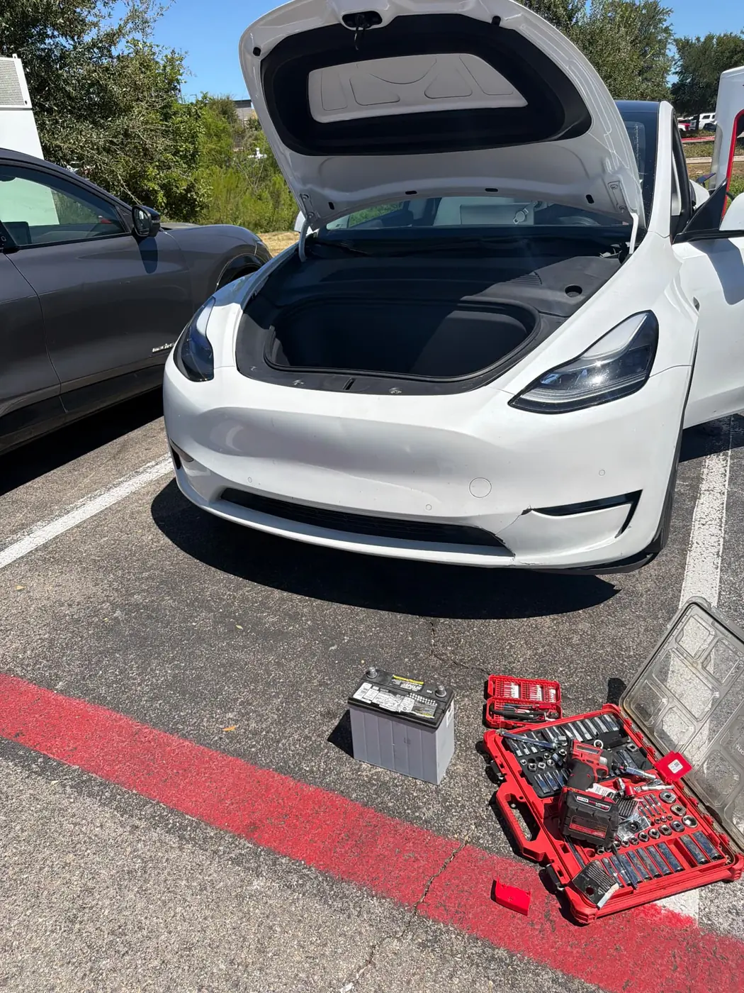 White Tesla Model Y with the frunk open in a Texas parking lot, the removed 12V battery and a socket set staged on the pavement during a mobile battery replacement.