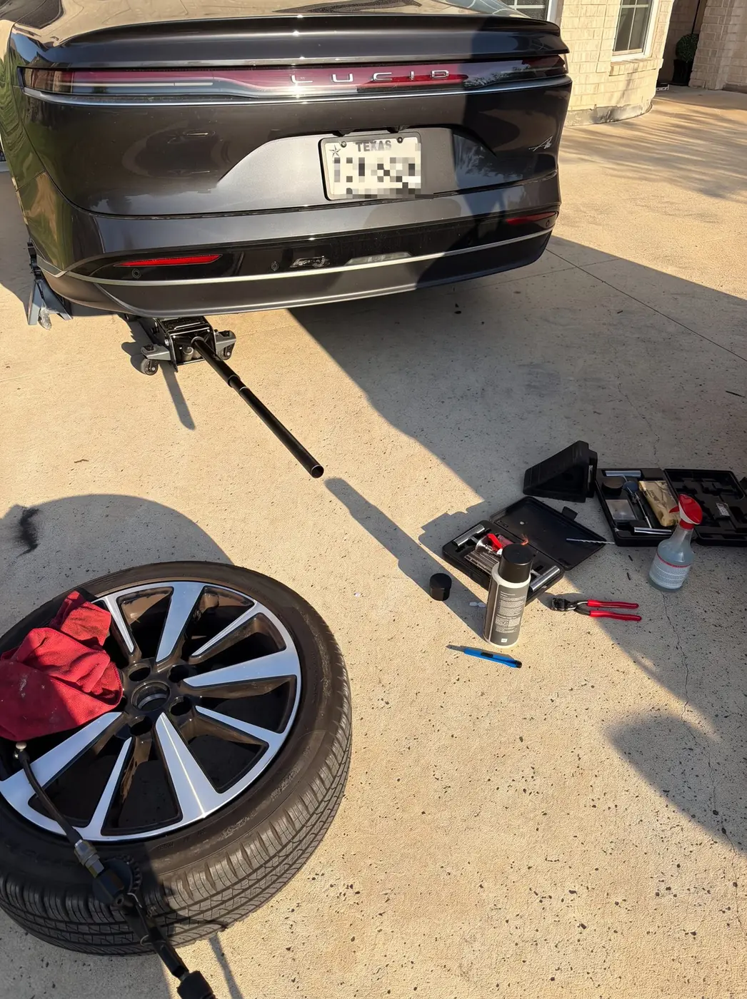 Lucid Air lifted on a floor jack in a Frisco, Texas driveway with the rear wheel removed and a tire plug kit staged for an on-site puncture repair.