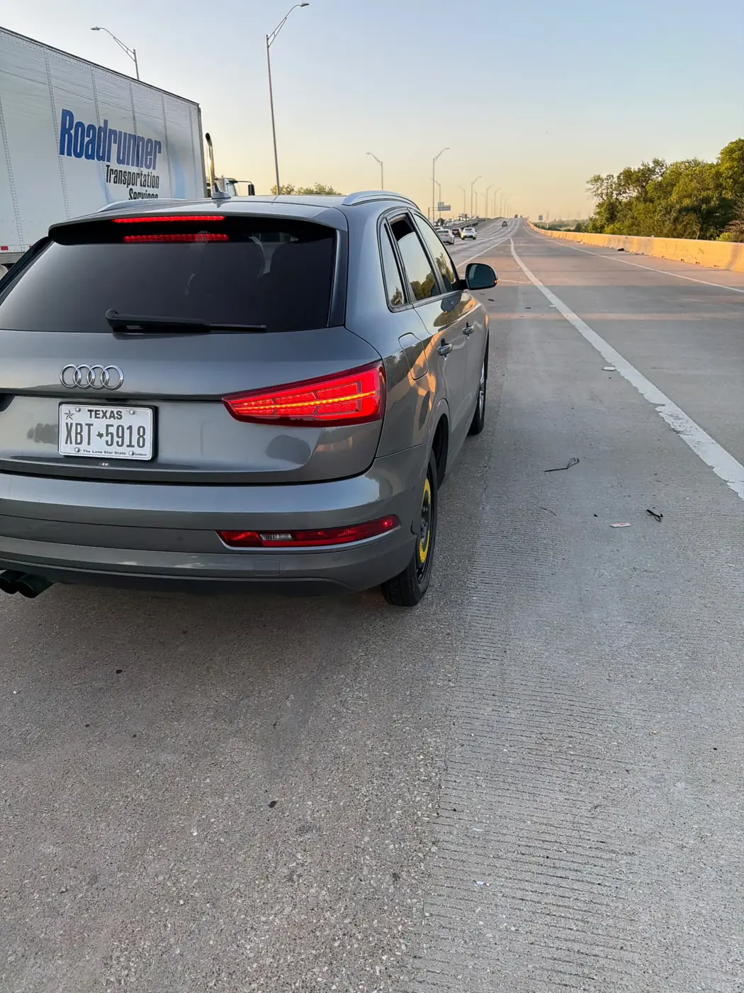 Grey Audi Q3 parked on the right shoulder of a Dallas-Fort Worth highway riding on a compact spare after a mobile flat tire change, semi-trailer passing on the left.