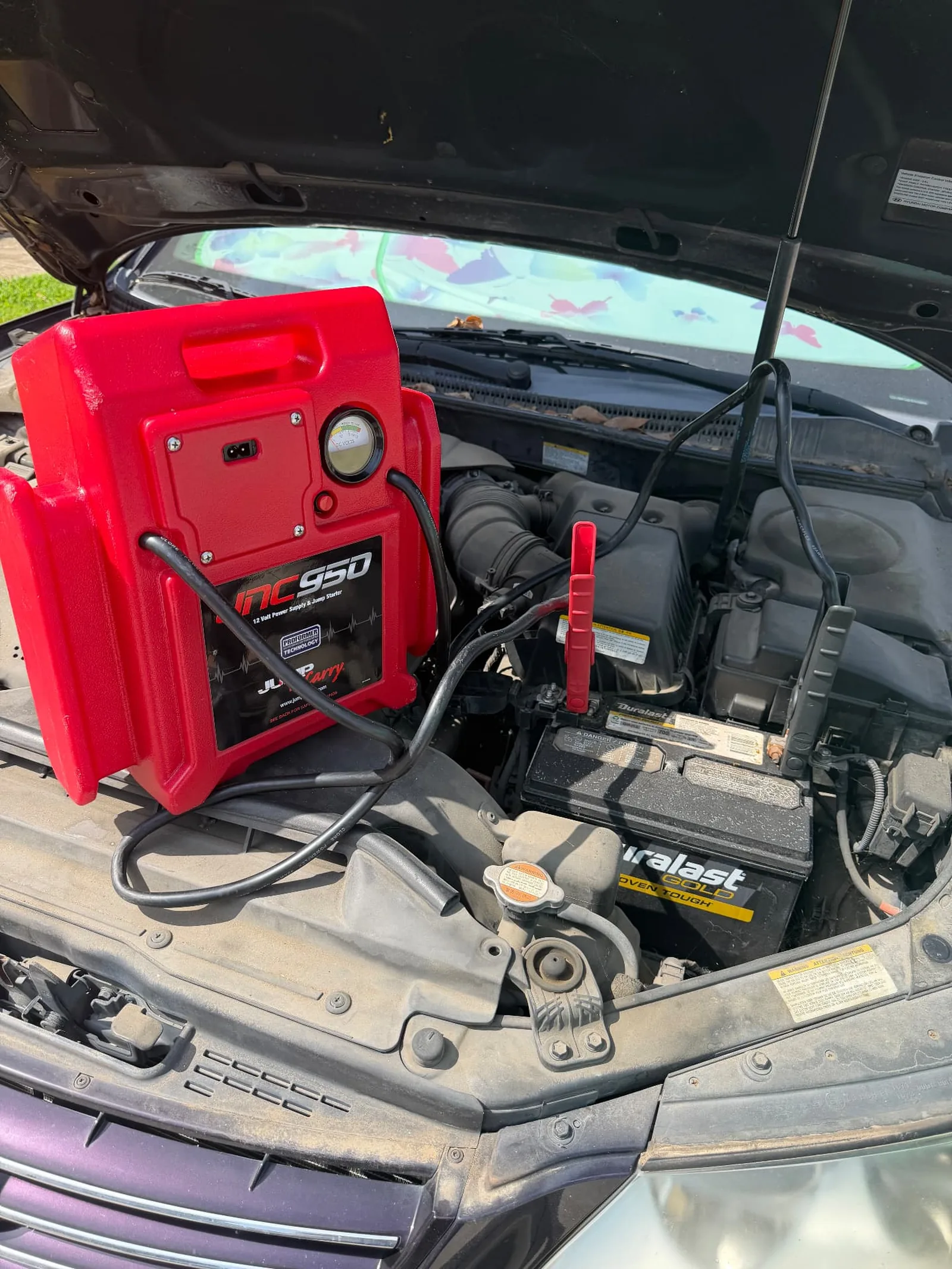 Red JNC950 jump pack sitting on the fender of a black Hyundai sedan in Frisco, Texas, with clamps on a Duralast Gold 12V battery during a mobile battery service call.