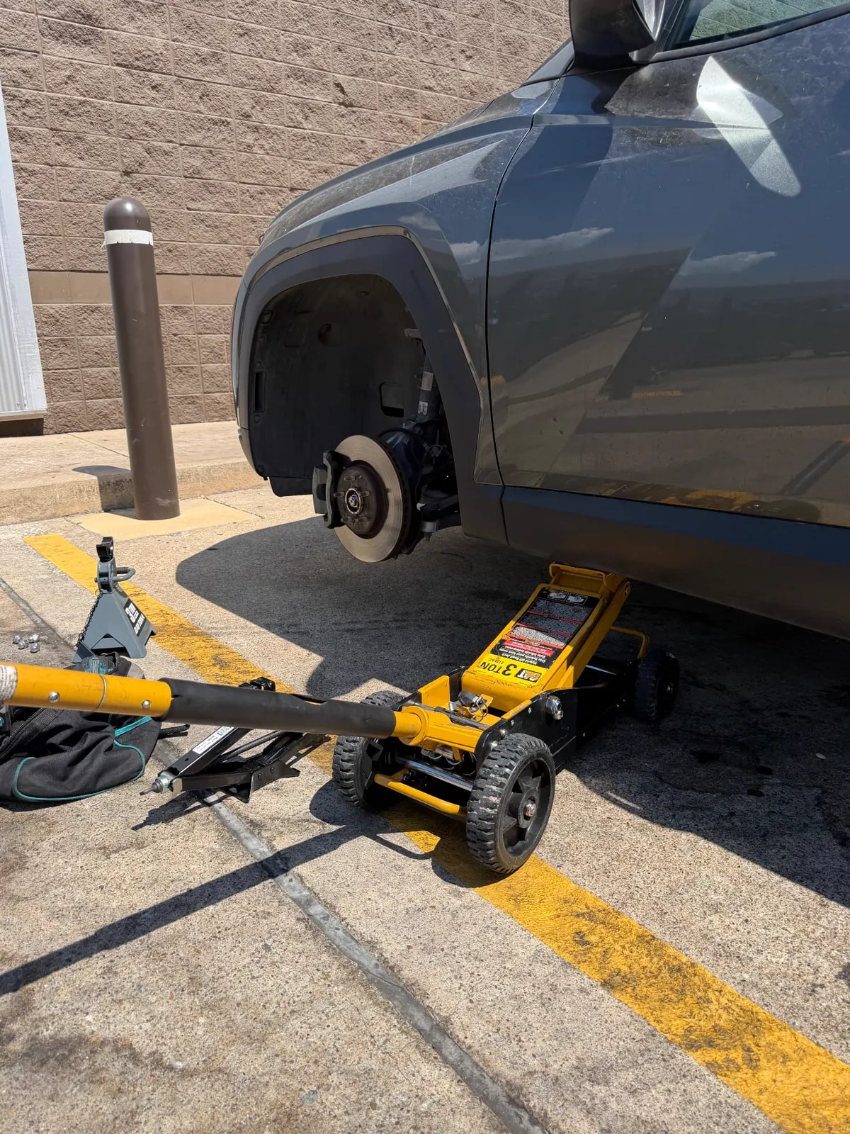 Grey SUV raised on a yellow three-ton floor jack with a jack stand in a McKinney, Texas parking lot, the front wheel removed and the brake rotor exposed during an on-site tire service call.
