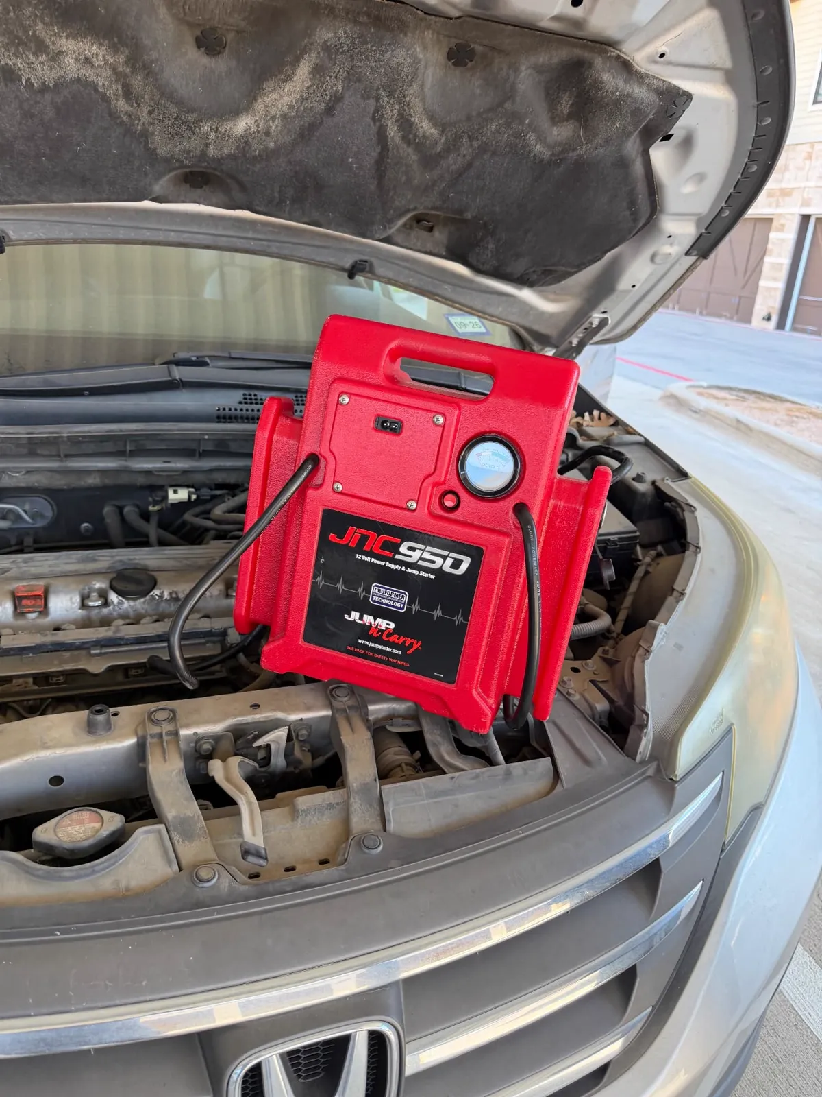 Red JNC950 12-volt jump pack resting on the radiator support of a silver Honda with the hood up outside a garage in Fort Worth, Texas, during a mobile jump start.