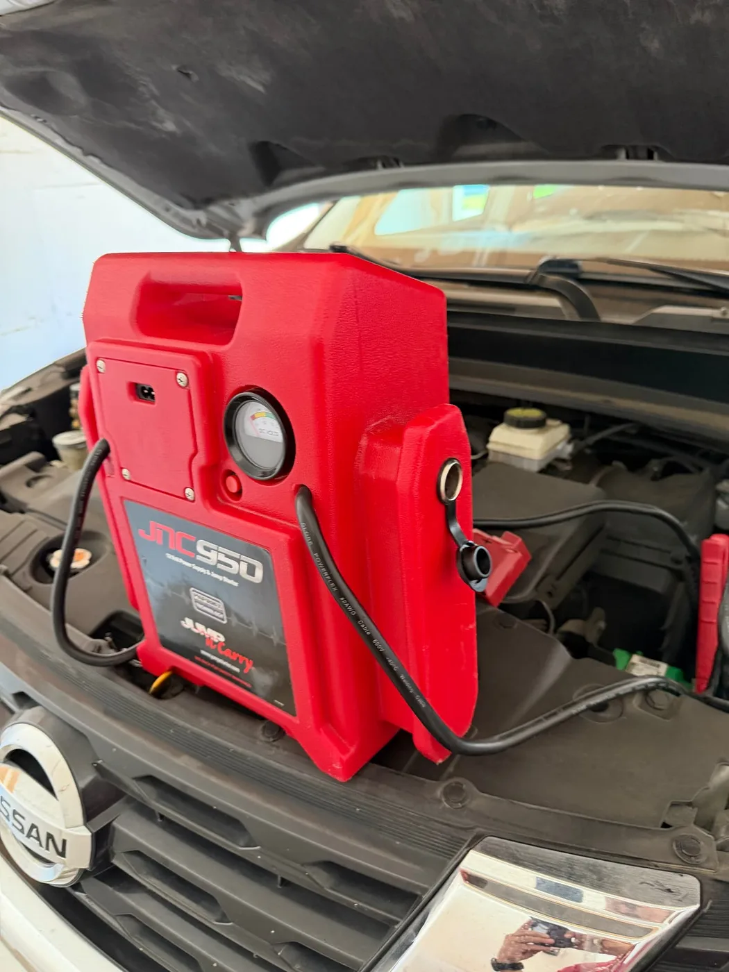 Red JNC950 commercial jump pack staged on the radiator support of a white Nissan with the hood up during a mobile jump start in Irving, Texas.