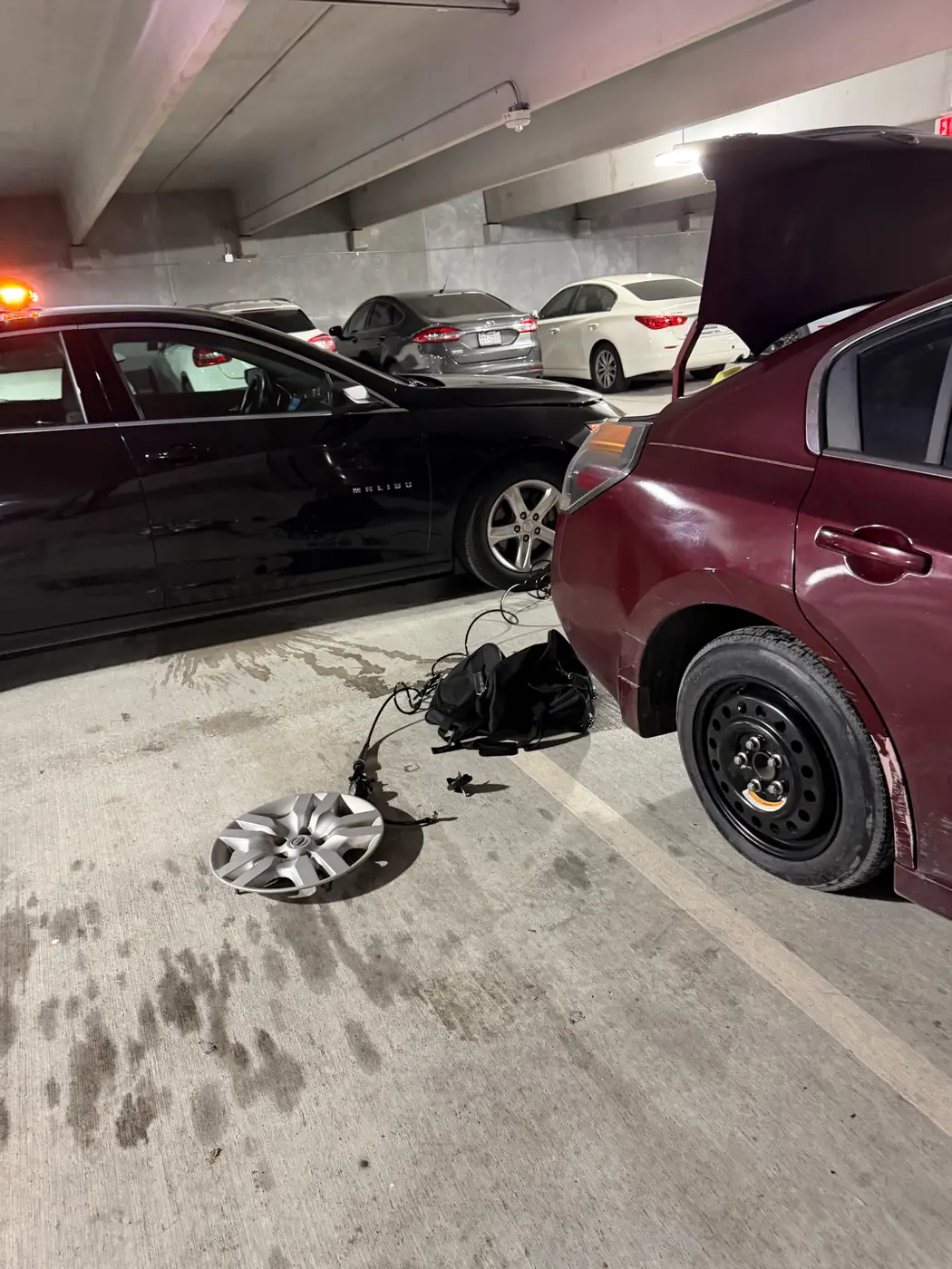 Nissan Altima on its spare wheel inside a Dallas parking garage after a late-night mobile tire change, hubcap and tool bag on the deck.