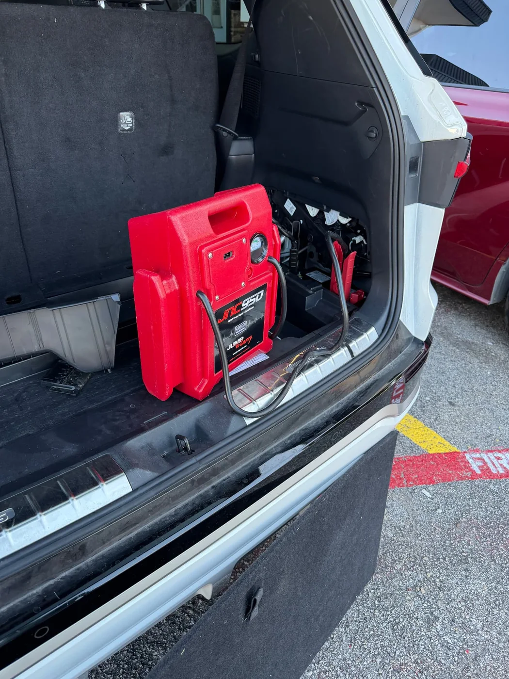 Red JNC950 commercial jump pack connected to the rear-mounted 12-volt battery of a white SUV during a mobile jump start in Dallas, Texas.