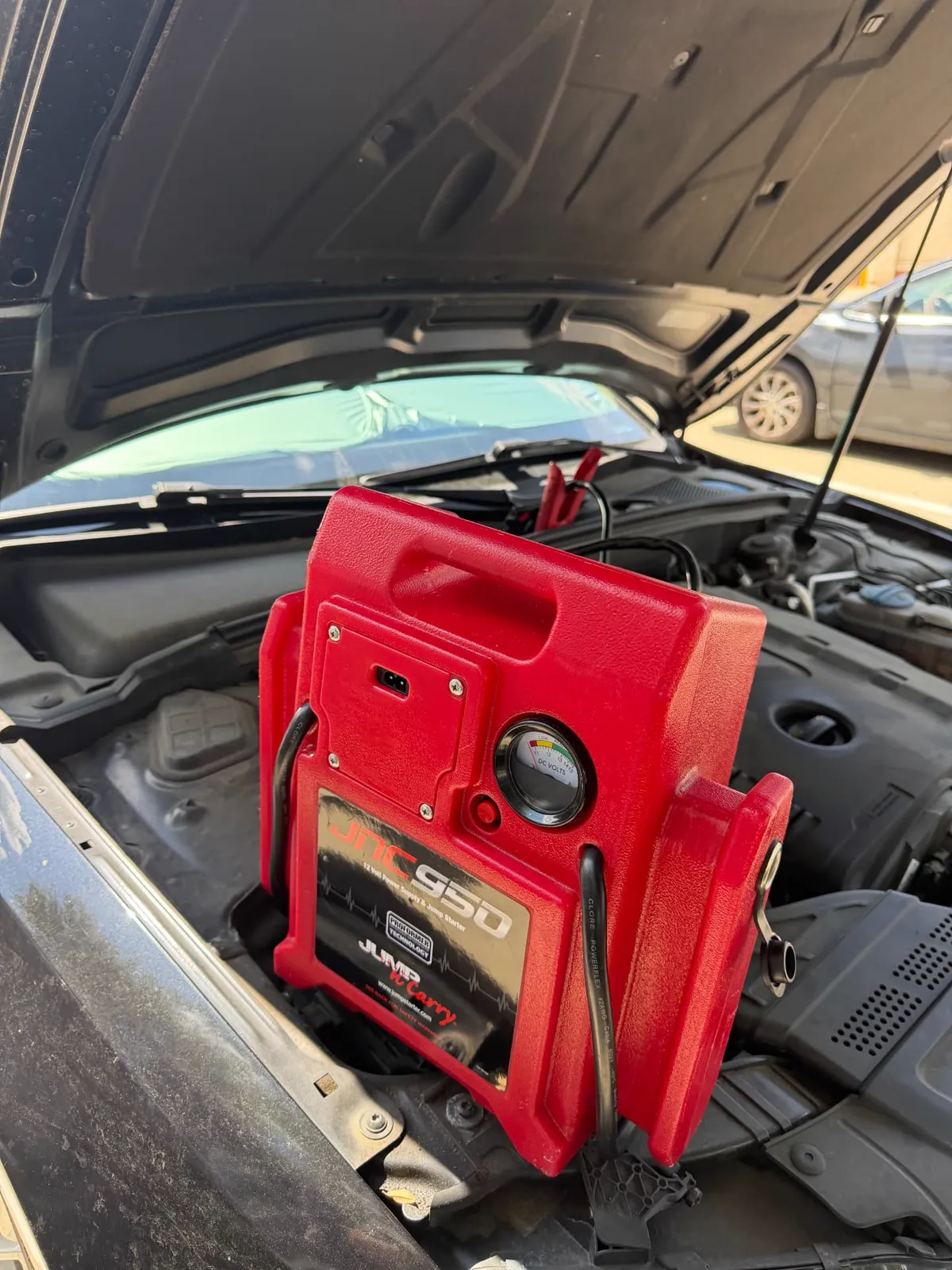 Red JNC950 12-volt jump pack sitting in the engine bay of a black sedan with the hood up in a Plano, Texas parking lot during a mobile jump start.